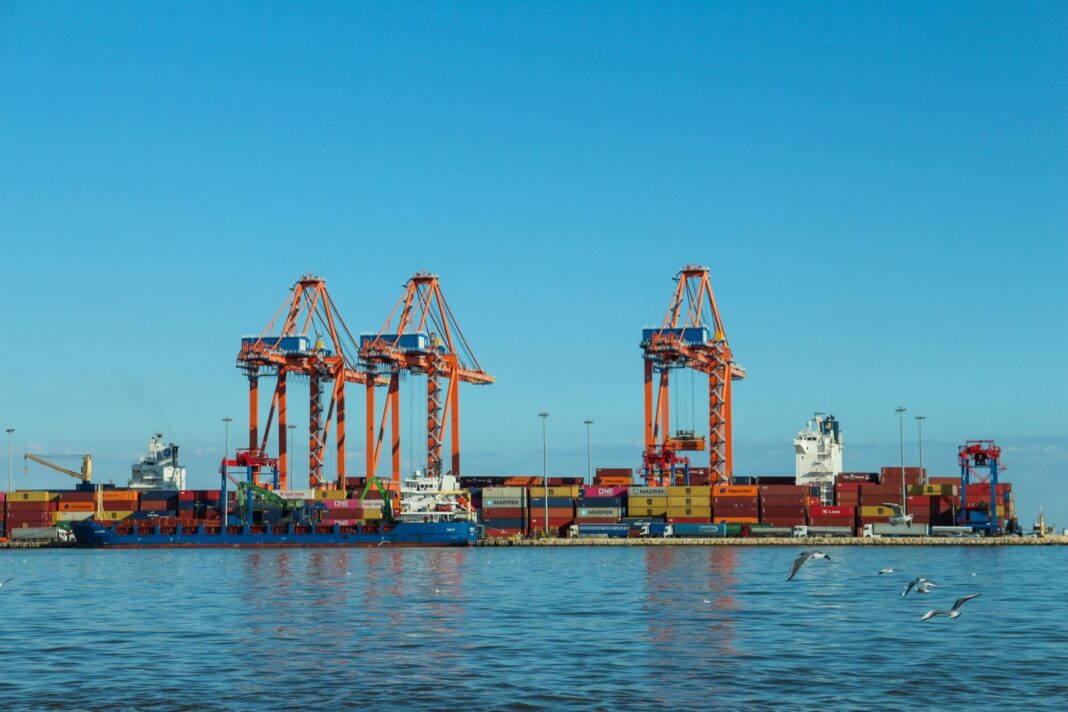 Grúas pórtico y contenedores apilados en un muelle con barcos atracados
