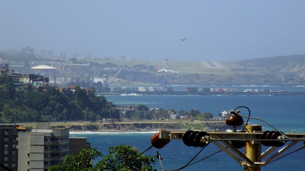 Vista del puerto de La Guaira con contenedores y grúas, y la pista del aeropuerto de Maiquetía al fondo