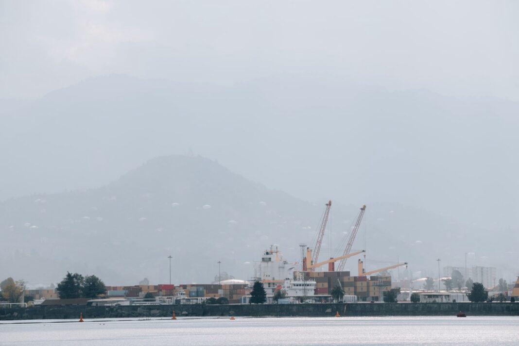 Puerto con grúas y contenedores bajo la niebla, con cerros al fondo