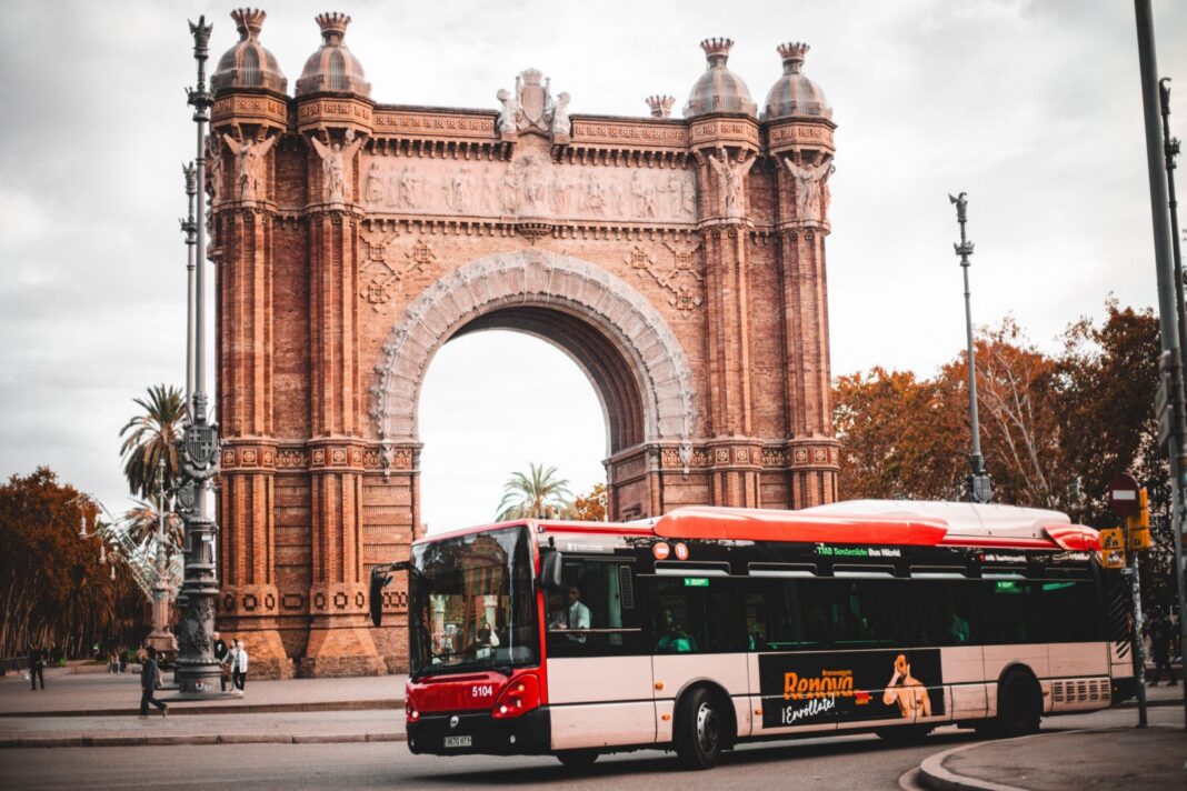 Autobús rojo y blanco de TMB pasa frente al Arco de Triunfo de Barcelona
