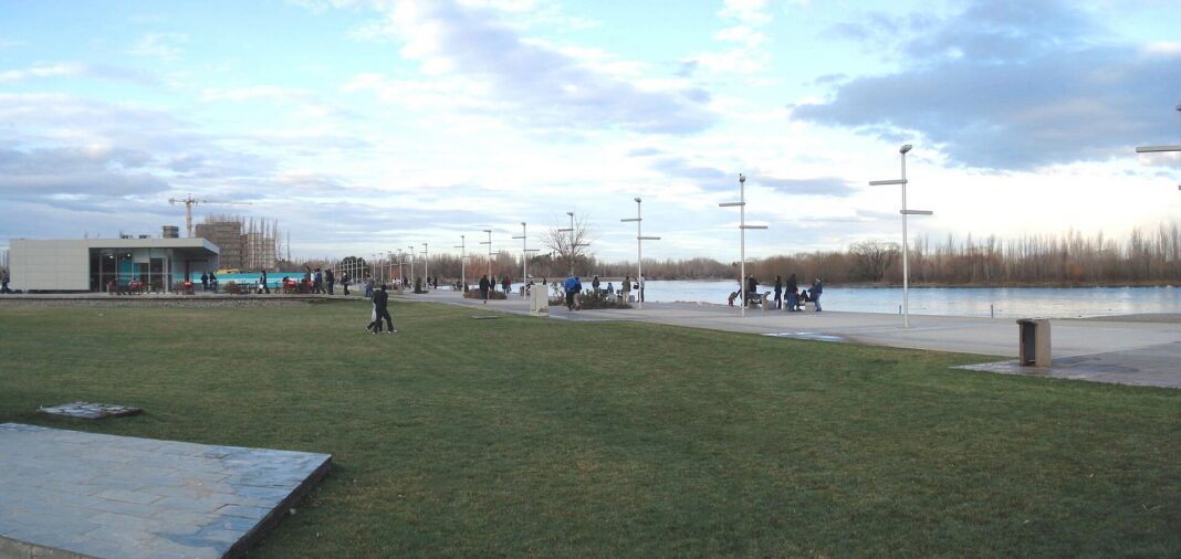Paseo costero junto al río Limay en Neuquén, con césped, faroles y gente caminando a la orilla