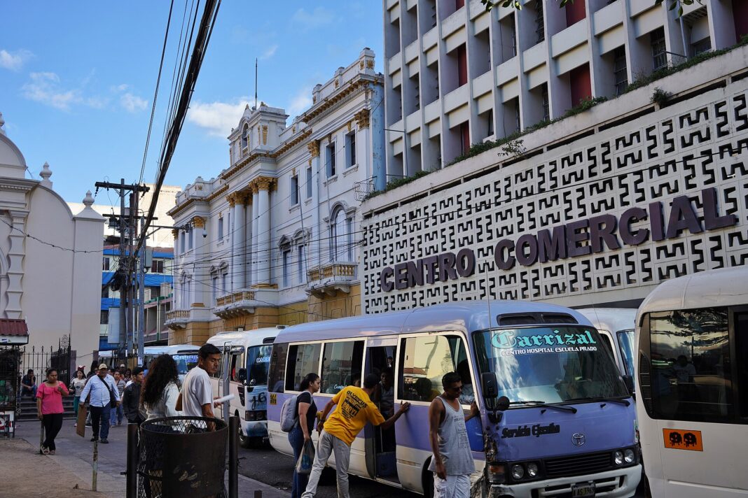 Microbuses de transporte urbano estacionados frente a un centro comercial en el centro de Tegucigalpa, con pasajeros subiendo