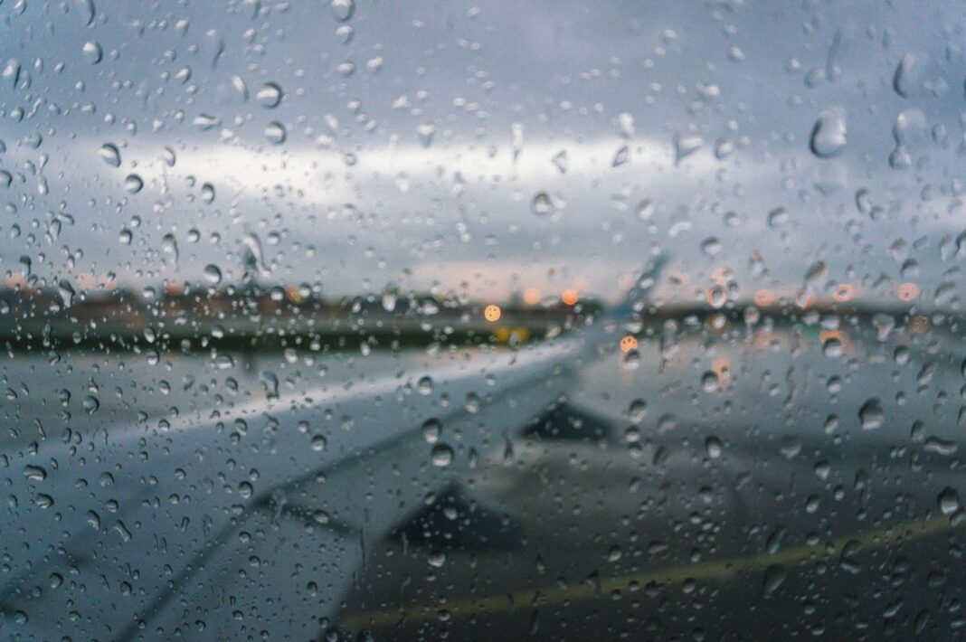 Ventanilla de avión cubierta de gotas de lluvia con el ala y la pista al fondo