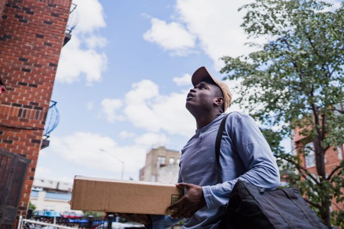 Las gafas de Amazon fotografiarán rutas de 5.000 repartidores Repartidor con una caja busca la dirección frente a un edificio de departamentos