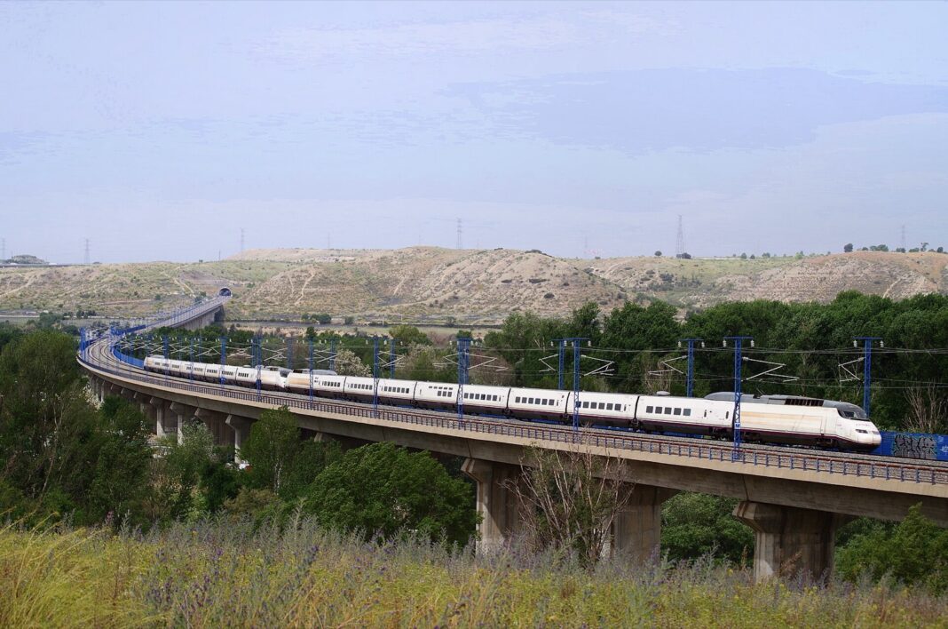 Dos trenes AVE de la serie 100 acoplados cruzan un viaducto de la línea de alta velocidad entre cerros y arboleda