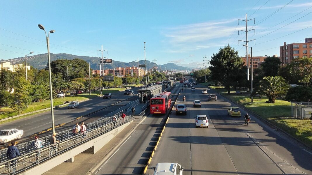 Autopista Norte de Bogotá vista desde un puente peatonal, con un bus rojo de TransMilenio en su carril exclusivo, autos y los cerros orientales al fondo