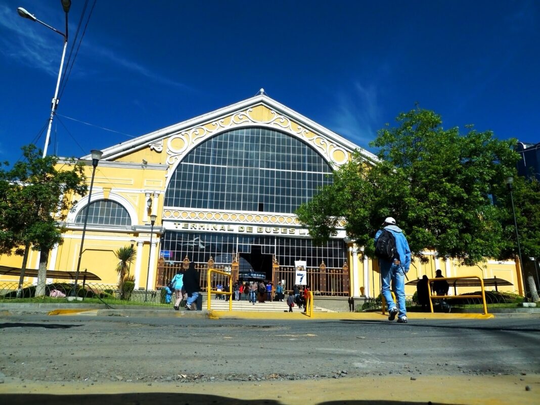 Fachada amarilla de la terminal de buses de La Paz con pasajeros entrando por la mañana bajo un cielo azul