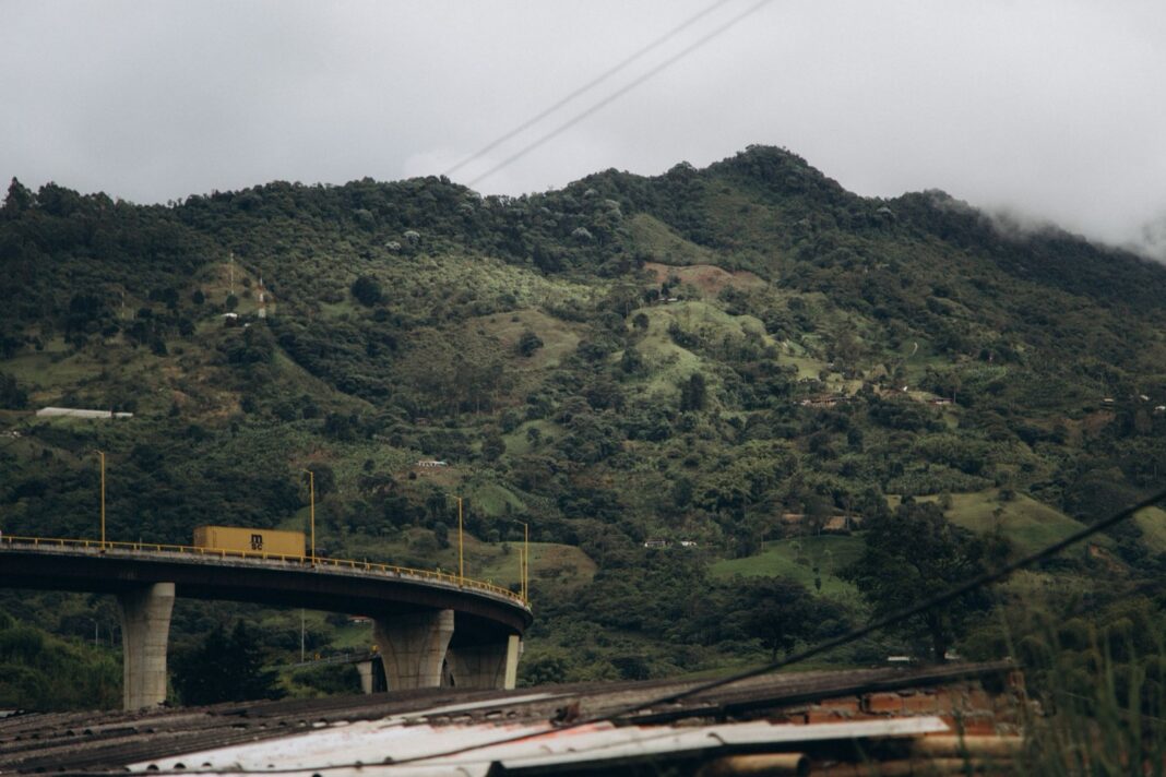 Tractocamión con contenedor cruza un viaducto de una carretera de montaña en Colombia, con laderas verdes y cielo nublado