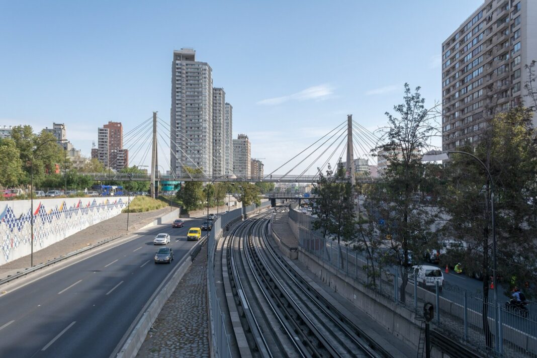 Autos circulan por la Autopista Central de Santiago junto a las vías del metro, con un puente atirantado al fondo