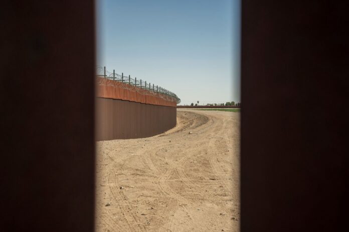 EE.UU. prohíbe drones en la frontera con México por un año Muro fronterizo de acero con alambre de púas junto a un camino de tierra, visto entre dos barrotes del propio muro
