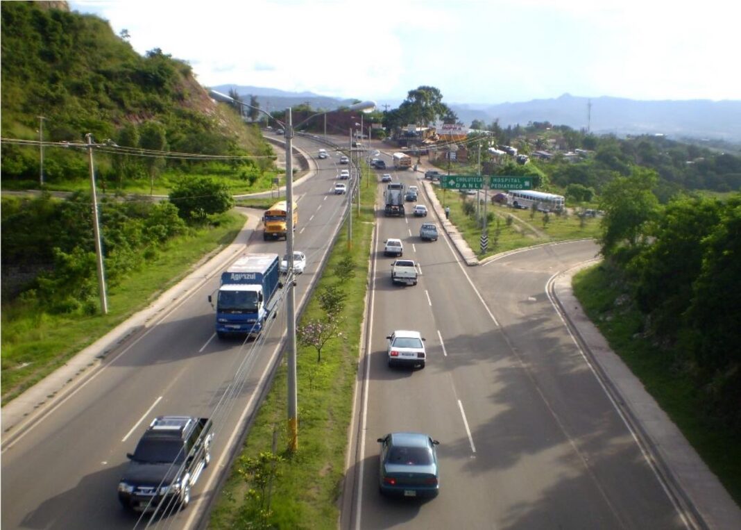 Tráfico de camiones, buses y autos en el Anillo Periférico de Tegucigalpa, con cerros verdes al fondo y señales hacia Choluteca y Danlí