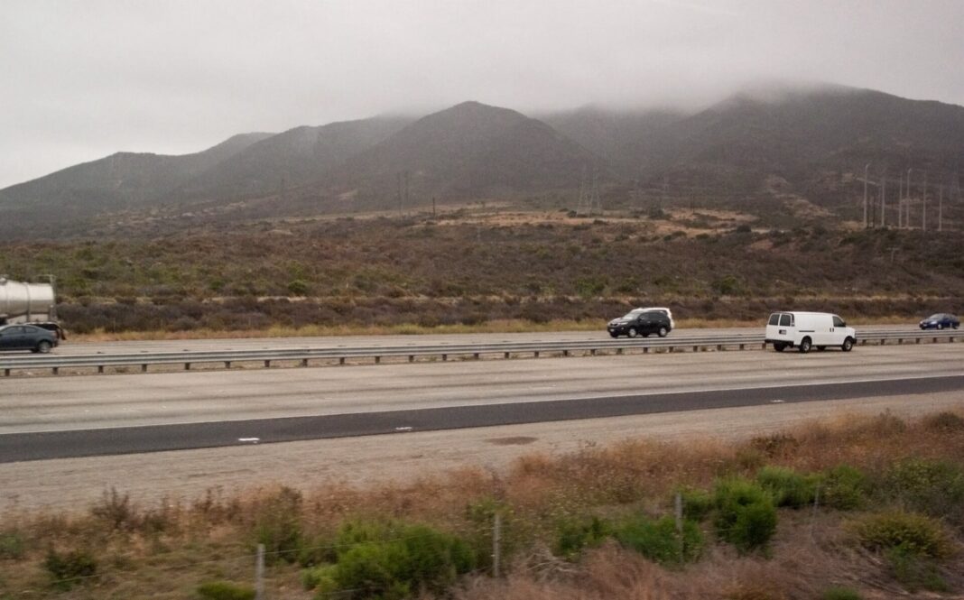 Interestatal 5 a su paso por Camp Pendleton, con un camión cisterna y vehículos ligeros bajo un cielo nublado y los cerros de la base al fondo