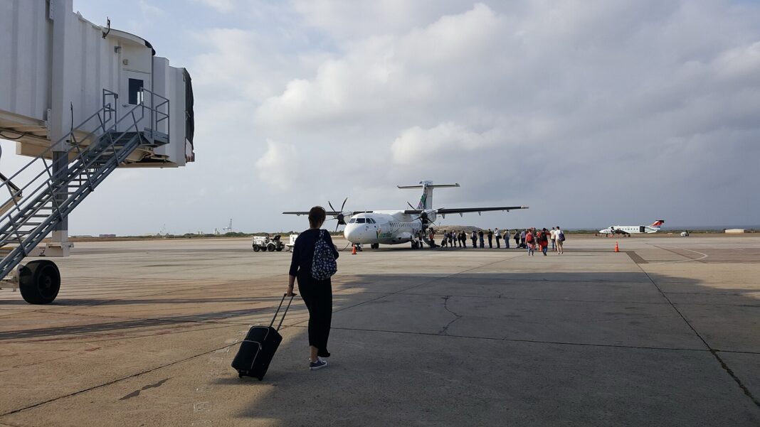 Pasajeros caminan por la plataforma del aeropuerto Reina Beatrix de Aruba hacia un turbohélice estacionado, con un puente de embarque a la izquierda