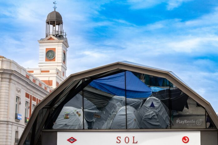 Metro de Madrid reabre la estación de Sol tras 33 horas cerrada Acceso acristalado de la estación de Sol en la Puerta del Sol de Madrid, con las tiendas de la acampada reflejadas en el vidrio y el reloj de la Casa de Correos al fondo