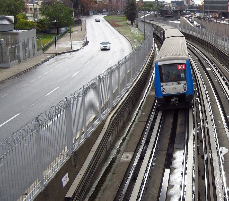 Tren de la Línea 2 del Metro de Santiago en superficie sobre vías mojadas, junto a una avenida con pavimento húmedo