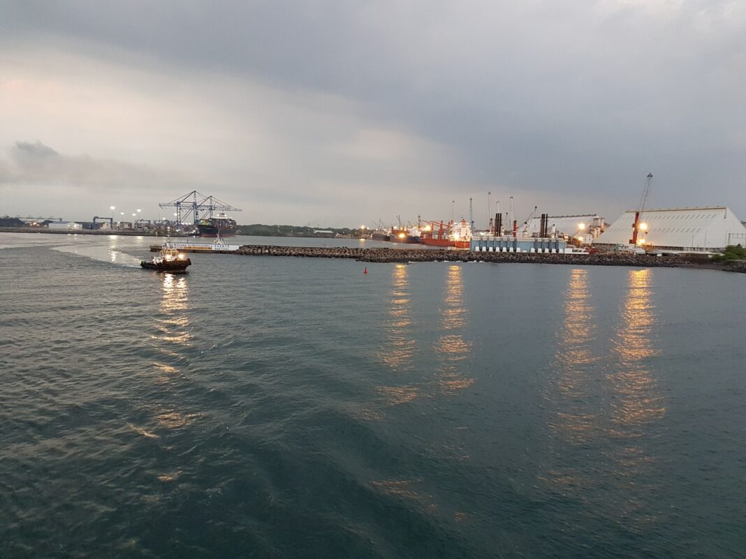 Muelles de Puerto Quetzal al anochecer, con grúas pórtico, buques atracados y un remolcador frente al letrero de bienvenida