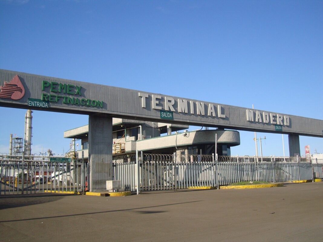 Acceso a la Terminal Madero de Pemex Refinación, con la caseta de control, la reja de entrada y las torres de la refinería al fondo bajo un cielo despejado