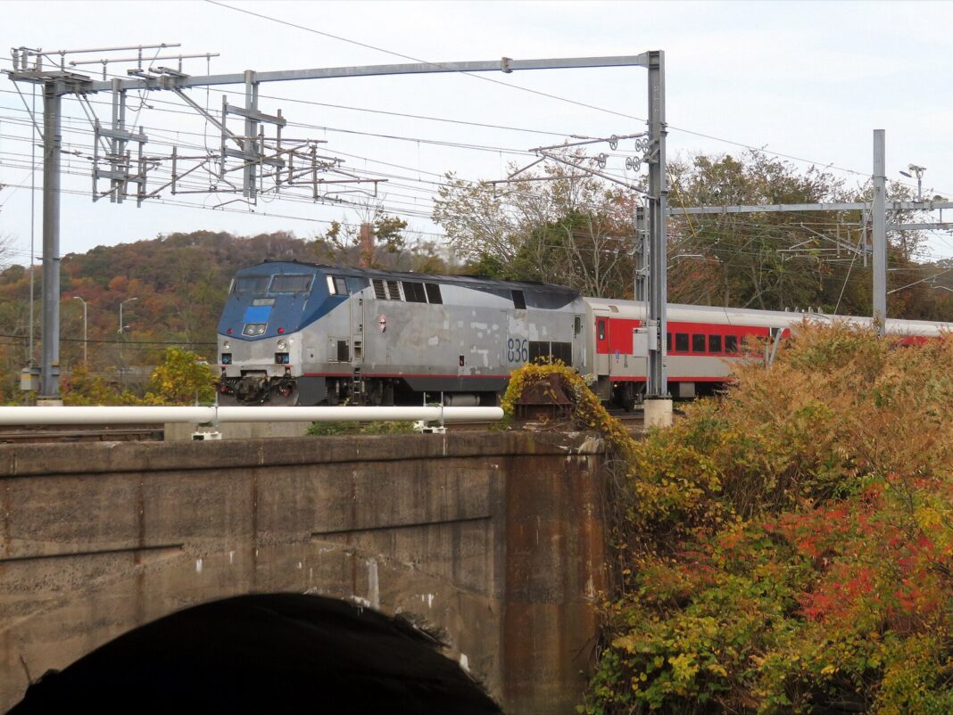 Tren de pasajeros con locomotora plateada y vagones rojos cruza un puente de piedra entre árboles otoñales